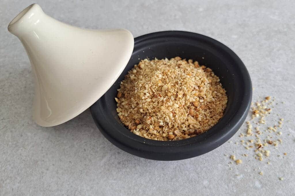 Pot with a lid open containing dukkah with some sprinkled on the work surface