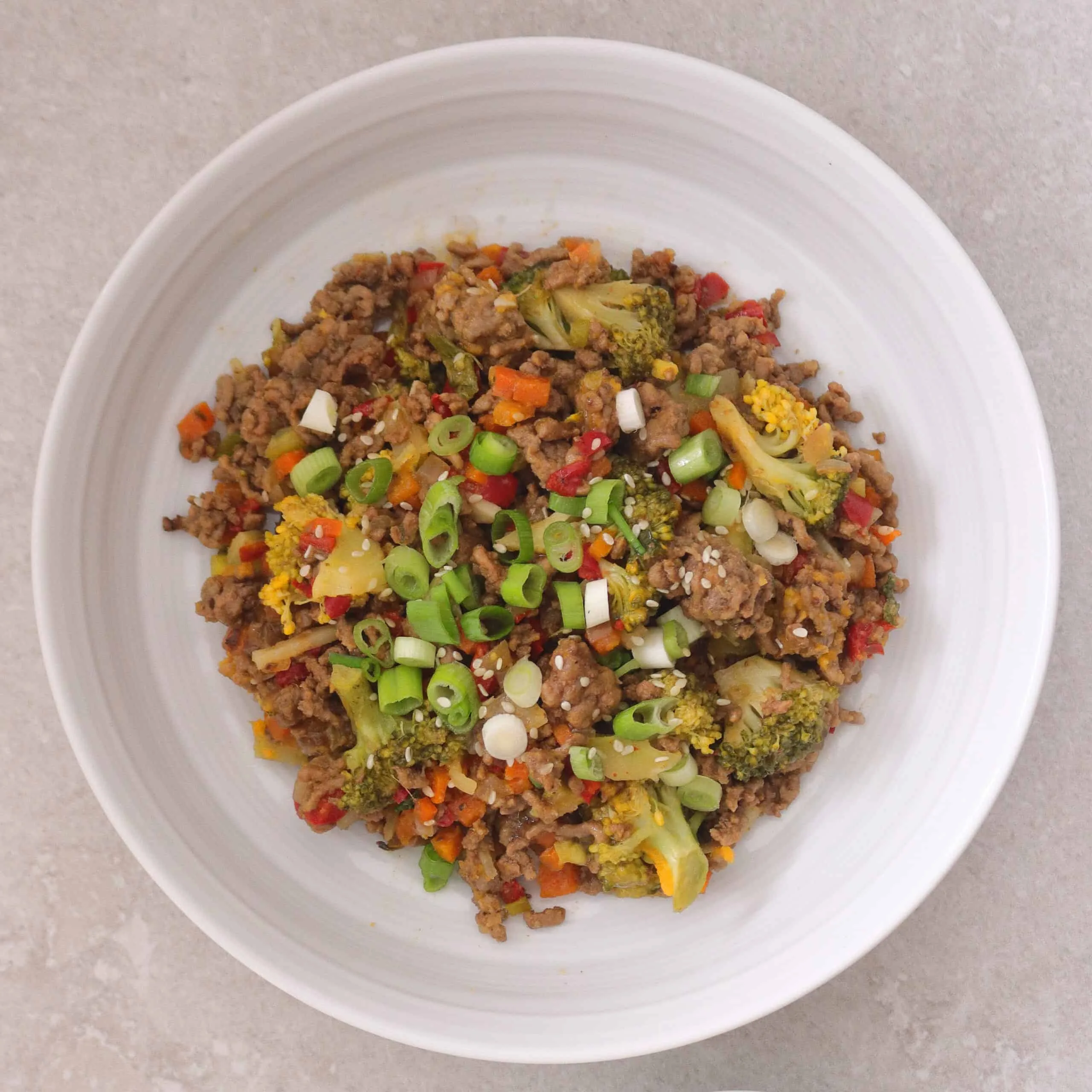 Bowl of beef mince stir fry with diced vegetables and broccoli sprinkle with sliced spring onion and sesame seeds.