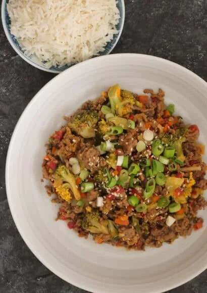 White bowl full of beef mince stir fry with diced vegetable, broccoli, and topped with sliced spring onion and sesame seeds on a dark board background alongside a small bowl of cooked rice