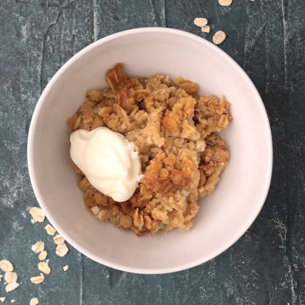 bowl of apple crumble with creme fraiche on a blue board with some loose oats