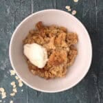 bowl of apple crumble with creme fraiche on a blue board with some loose oats