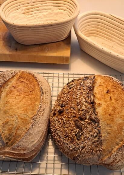 2 baked sourdough loaves cooling on a wire rack in front of the baking bread baskets