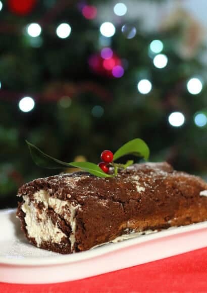 A beautiful, realistic Yule log cake (Bûche de Noël) resting on a festive ceramic plate. The cake is decorated with realistic chocolate bark texture, a dusting of powdered sugar 'snow', and topped with vibrant green holly leaves and bright red berries. In the background, a Christmas tree with soft, twinkling warm lights is slightly out of focus, creating a cozy, festive bokeh effect. The lighting is warm and inviting.