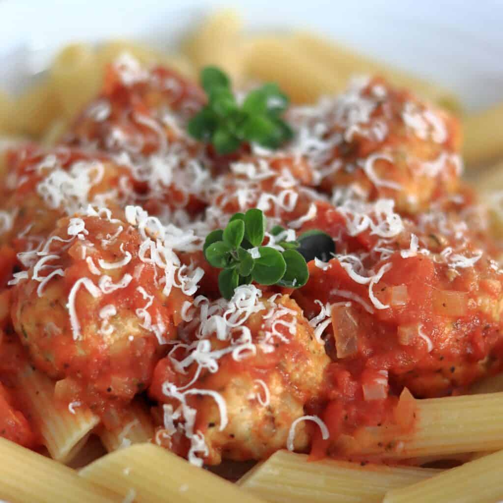 Bowl of pasta topped with chunky tomato sauce and chicken meatballs with a sprinkling of parmesan and a couple of sprigs of oregano