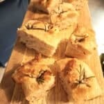 sliced squares of focaccia bread topped with rosemary and sprinkled with sea salt on a long wooden board