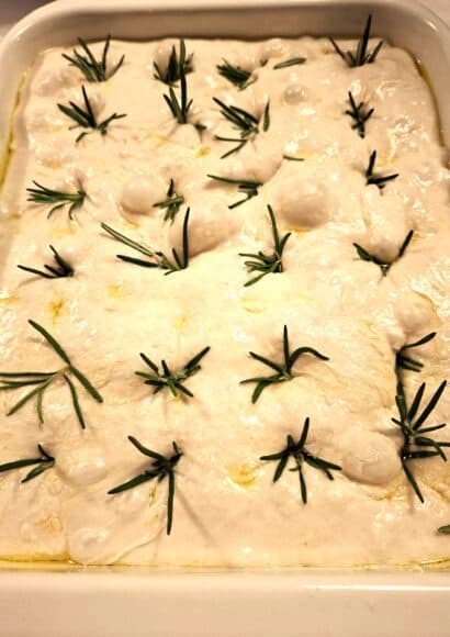 a tray full of unbaked sourdough focaccia, studded with rosemary springs and sprinkled with sea salt