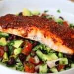 A close up of a Cajun-spiced salmon fillet resting on a vibrant Mexican-inspired salad with tomato, cucumber,, black beans, red onion, red chilli, coriander, and lime dressing