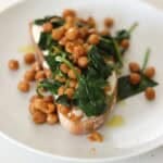 A slice of toast on a white plate onto which is spread a generous helping of whipped feta. On top of this are smoky cooked chickpeas and spinach which has all been drizzled with a little olive oil.