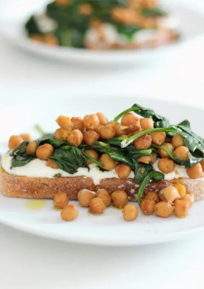 A white plate in the foreground and on it is a slice of thick toast, topped with creamy feta, then a mix of chickpeas and wilted spinach