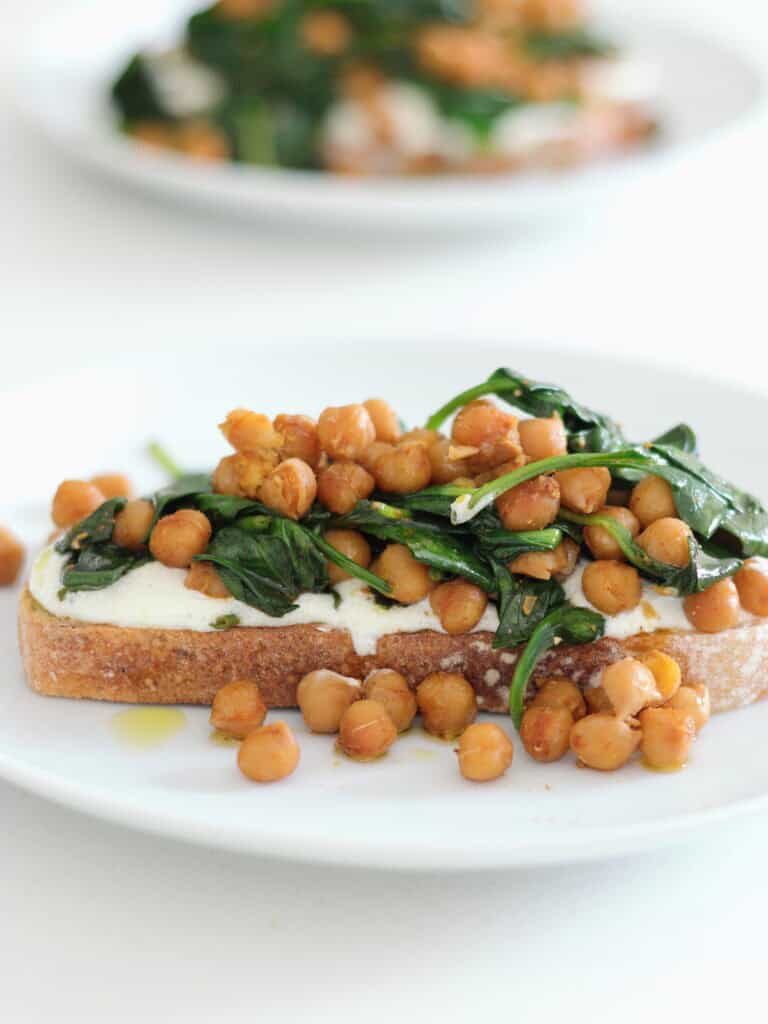 A white plate in the foreground and on it is a slice of thick toast, topped with creamy feta, then a mix of chickpeas and wilted spinach