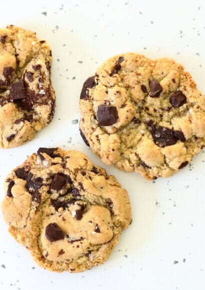 A close up of 3 chocolate chip cookies on a worktop, slightly golden at the edges, with plenty of molten chocolate chunks.