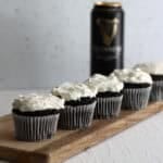 A line of dark chocolate muffins in their cases, topped with a thick pile of creamy icing, sat on a wooden board infront of a can of Guinness