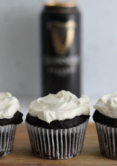 Three dark chocolate muffins, topped thickly with cream cheese icing, lined up in a row facing front on a board in front of a can of Guinness