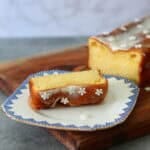 A slice of rectangular lemon cake that is topped with lemon drizzle icing and some white icing flowers. Sat in front of the remaining loaf cake of lemon drizzle on a wooden chopping board.