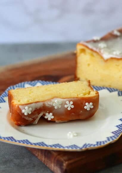 A slice of rectangular lemon cake that is topped with lemon drizzle icing and some white icing flowers. Sat in front of the remaining loaf cake of lemon drizzle on a wooden chopping board.