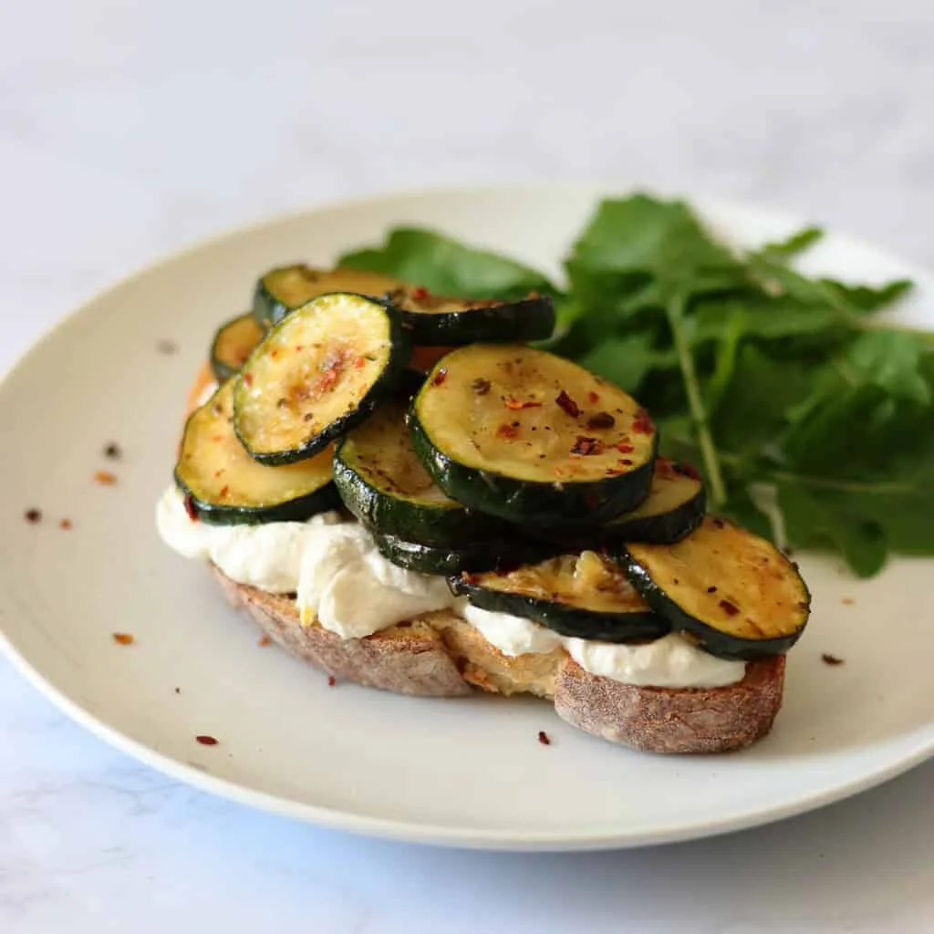 A plate with a slice of sourdough toast, topped with white creamy ricotta and then piled high with slow-cooked courgettes flecked with fennel and chilli flakes