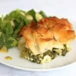 Plate holding a slice of Mediterranean courgette and feta pie, with spinach, topped with golden brown crispy scrunched up filo pastry and served with a green salad and dressing.