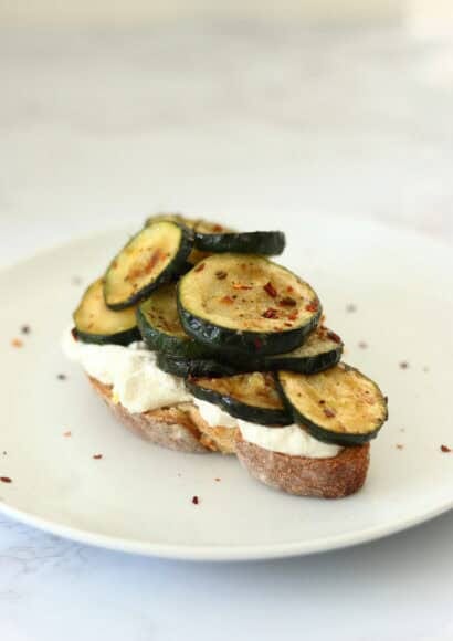A slice of sourdough toast, centred on a plate, topped with creamy white ricotta and piled high with soft and sticky courgette slices sprinkled with chilli flakes