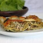 A white plate with a slice of lasagne showing the layers of courgette, pesto and walnut, topped with a crispy cheese layer and sage leaves. In front of a wooden bowl full of crunchy lettuce leaves