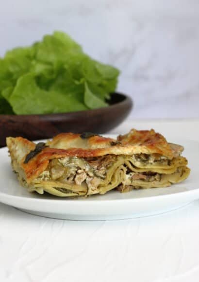 A white plate with a slice of lasagne showing the layers of courgette, pesto and walnut, topped with a crispy cheese layer and sage leaves. In front of a wooden bowl full of crunchy lettuce leaves