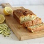 A golden coloured courgette lemon and poppy seed loaf cake on a rectangular board, with 2 slices cut showing the inside full of courgettes and poppy seeds. Beside the board is some grated courgette and a lemon sliced in half.