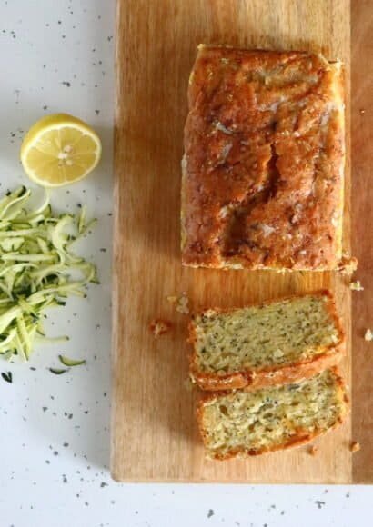 A golden coloured courgette lemon and poppy seed loaf cake on a rectangular board, with 2 slices cut showing the inside full of courgettes and poppy seeds. Beside the board is some grated courgette and a lemon sliced in half.