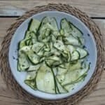 A white bowl placed on a raffia placemat on a wooden table. The bowl is full of thin strips of courgette, in a simple dressing with chopped mint and sprinkled with chilli flakes