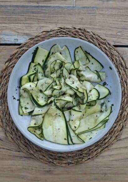 A white bowl placed on a raffia placemat on a wooden table. The bowl is full of thin strips of courgette, in a simple dressing with chopped mint and sprinkled with chilli flakes