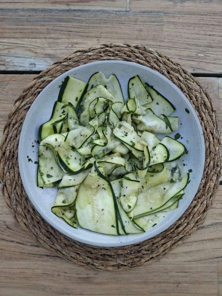 A white bowl placed on a raffia placemat on a wooden table. The bowl is full of thin strips of courgette, in a simple dressing with chopped mint and sprinkled with chilli flakes