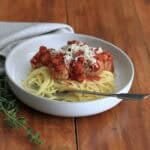 A cream bowl full of spaghetti topped with turkey and courgette meatballs in tomato sauce, on a wooden table. The bowl has a fork in it, beside the bowl is some fresh oregano and behind the bowl is a taupe coloured napkin