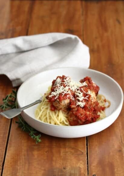 A cream bowl full of spaghetti topped with turkey and courgette meatballs in tomato sauce, on a wooden table. The bowl has a fork in it, beside the bowl is some fresh oregano and behind the bowl is a taupe coloured napkin