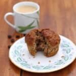 A courgette and carrot muffin on a plate, broken apart to show the threads of courgette and carrot inside the moist sponge, sat in front of a cup of coffee