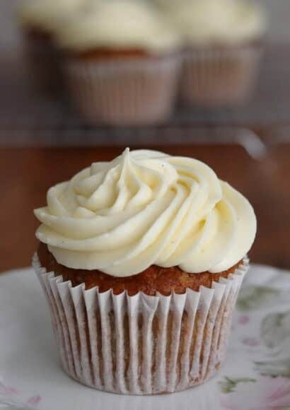 Close up of an iced courgette and carrot muffin, in its paper case on a plate in the foreground, with more iced muffins in the background on a wire rack