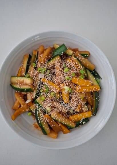 A medium white bowl filled with batons of green and yellow courgette that have been stir-fried in a sauce of soy, hoisin, and sprinkled with sesame seeds.