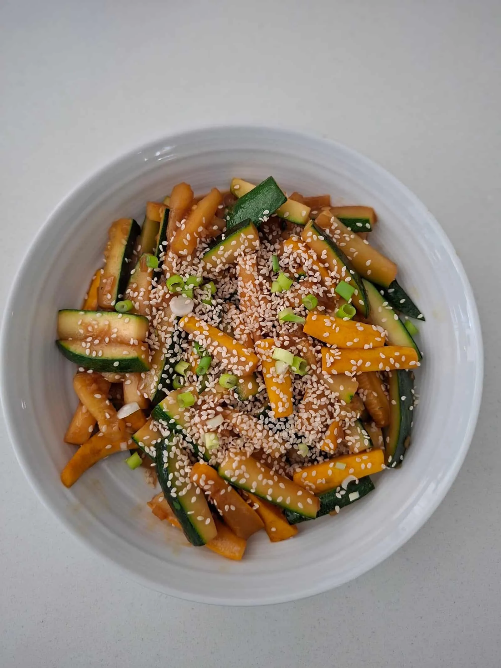 A medium white bowl filled with batons of green and yellow courgette that have been stir-fried in a sauce of soy, hoisin, and sprinkled with sesame seeds.