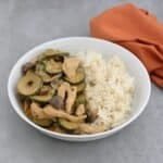 A white bowl, on a grey countertop, two thirds filled with strips of chicken, sliced courgette and quartered mushrooms all in a creamy paprika flavoured sauce, served with rice.