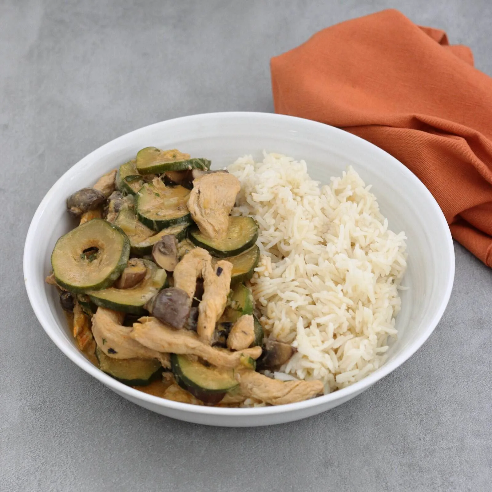 A white bowl, on a grey countertop, two thirds filled with strips of chicken, sliced courgette and quartered mushrooms all in a creamy paprika flavoured sauce, served with rice.