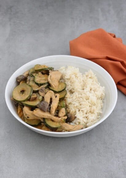 A white bowl, on a grey countertop, two thirds filled with strips of chicken, sliced courgette and quartered mushrooms all in a creamy paprika flavoured sauce, served with rice.