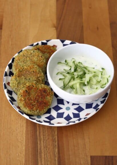 Fresh courgette and chickpea falafels arranged with on a colourful plate with a dipping bowl of tzatziki sauce topped with grated cucumber