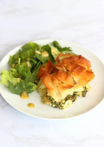 Plate holding a slice of Mediterranean courgette and feta pie, with spinach, topped with golden filo pastry beside a side salad of lettuce