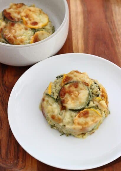 In the foreground is a single round serving of the gratin, showing the layers of grated courgette and thick sauce inside, topped with sliced green and yellow courgettes and melted cheese.