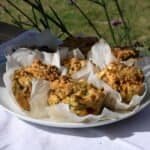 A white plate on a white tablecloth outside in the garden holding 6 golden courgette and feta savoury muffins in their paper cases