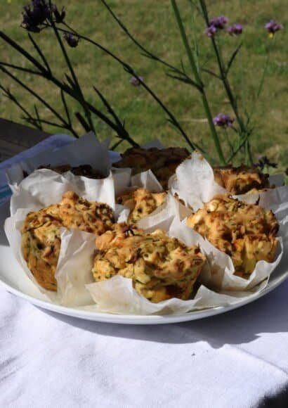 A white plate on a white tablecloth outside in the garden holding 6 golden courgette and feta savoury muffins in their paper cases