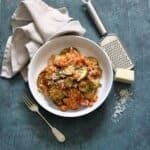 A white bowl on a blue board, full of a red tomato risotto and soft cooked slices of courgette sprinkle with cheese. In the background in a beige napkin, a fork and a grater with parmesan cheese scattered on the surface.