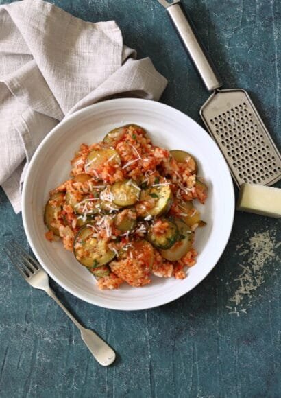 A white bowl on a blue board, full of a red tomato risotto and soft cooked slices of courgette sprinkle with cheese. In the background in a beige napkin, a fork and a grater with parmesan cheese scattered on the surface.