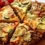 A wooden board, on top of which is a frittata cut into 6 pieces beside a bowl of salad leaves. The frittata is golden on top, and shows the slices of courgette and sweet potato inside.