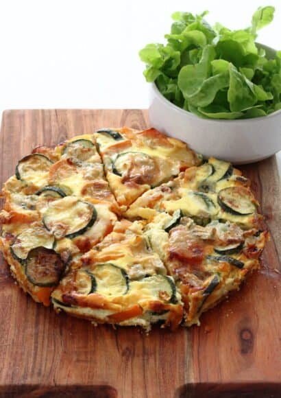 A wooden board, on top of which is a frittata cut into 6 pieces beside a bowl of salad leaves. The frittata is golden on top, and shows the slices of courgette and sweet potato inside.
