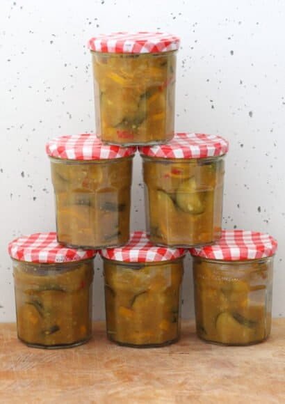 A pyramid of six jars filled with courgette chutney. The chutney shows visible courgette pieces, red chilli flecks and a glossy, tangy sauce with warm spices. The jars are stacked neatly, highlighting the texture and colour of the chutney