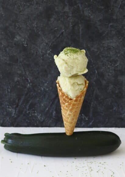 A waffle cone stood in a small hole cut into a courgette that is lying on a white worktop and a black background. The cone is filled with two scoops of light green ice cream on top of one another, sprinkled with fine lime zest on top.