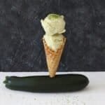 A waffle cone stood in a small hole cut into a courgette that is lying on a white worktop and a black background. The cone is filled with two scoops of light green ice cream on top of one another, sprinkled with fine lime zest on top.